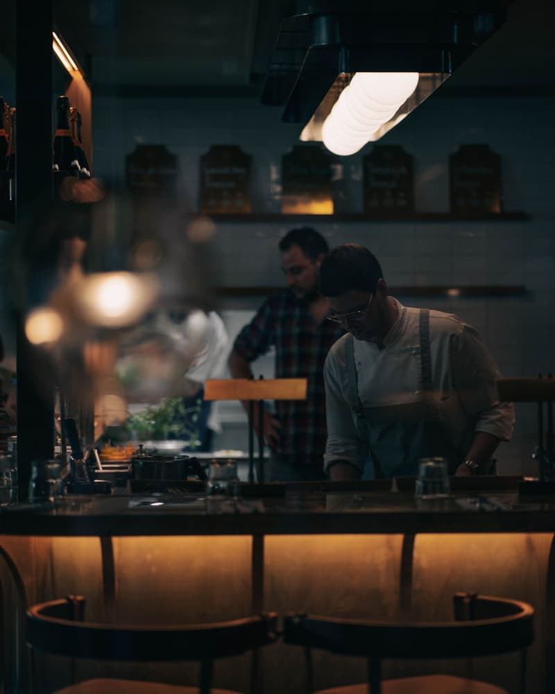Barra iluminada frente a la cocina abierta, con cocineros trabajando al fondo