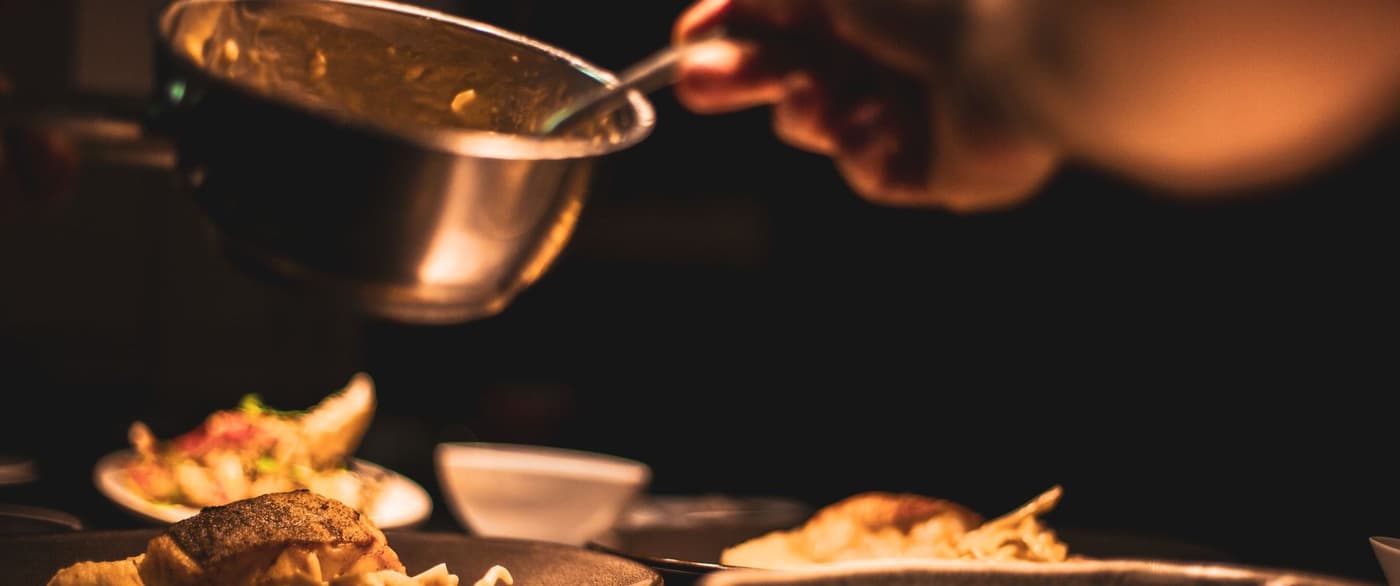 Mano del cocinero sirviendo salsa desde una olla de cobre sobre platos en penumbra