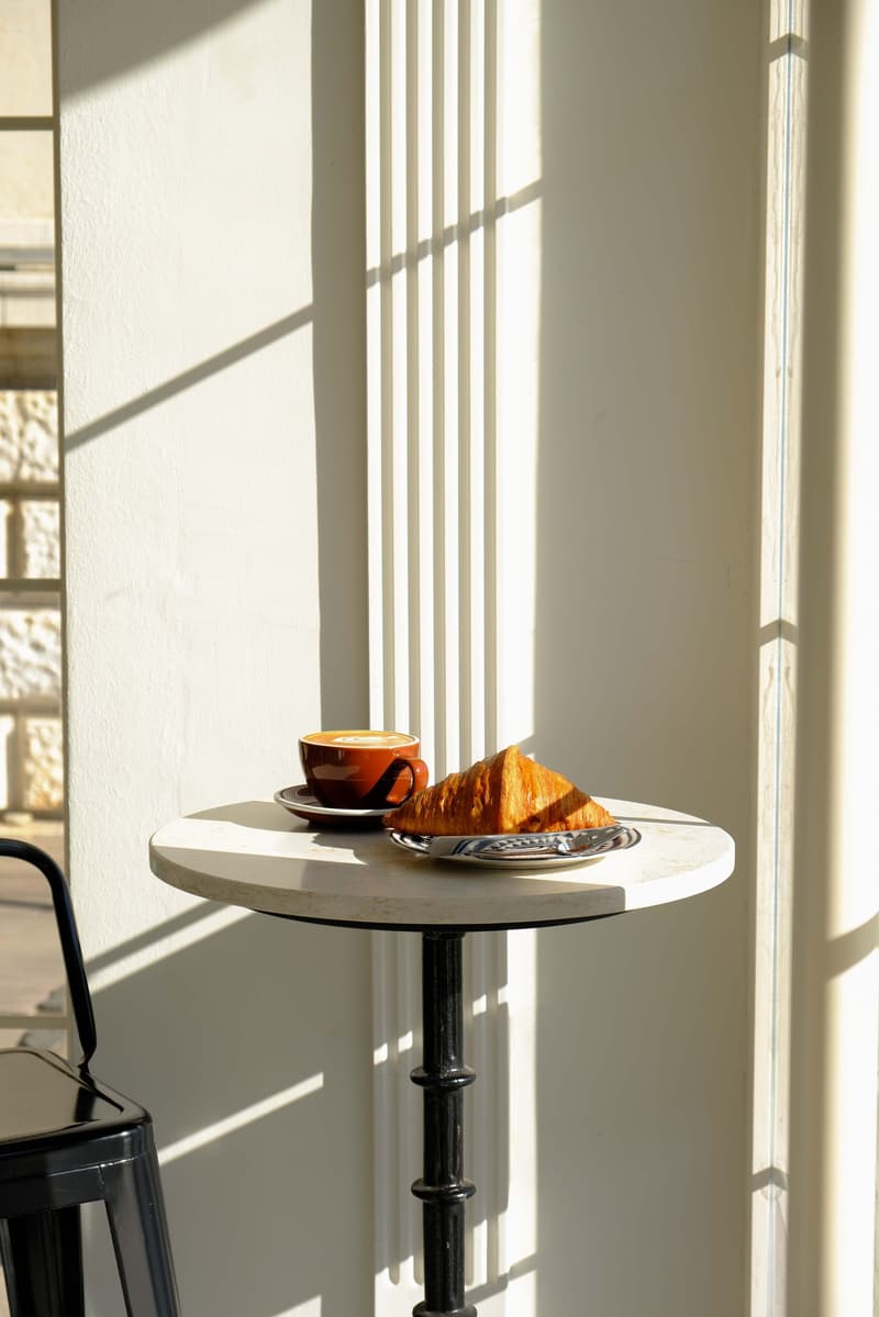 Mesa pequeña con café y pan junto a una ventana, bañada por el sol